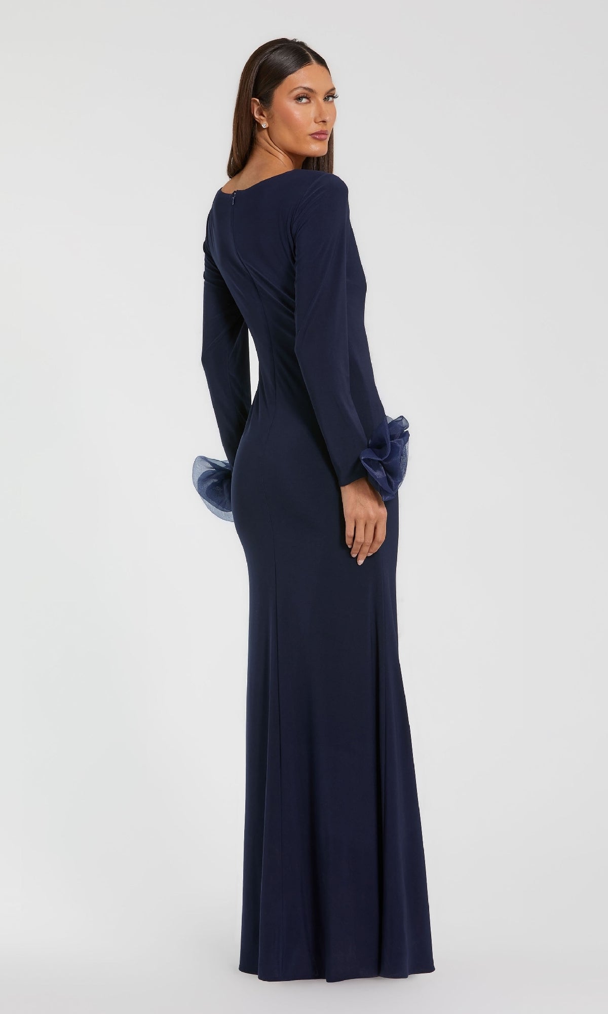 A woman stands facing away from the camera against a plain background, wearing the Mac Duggal 49949 long-sleeve formal dress—a floor-length navy gown with ruffled cuff details.